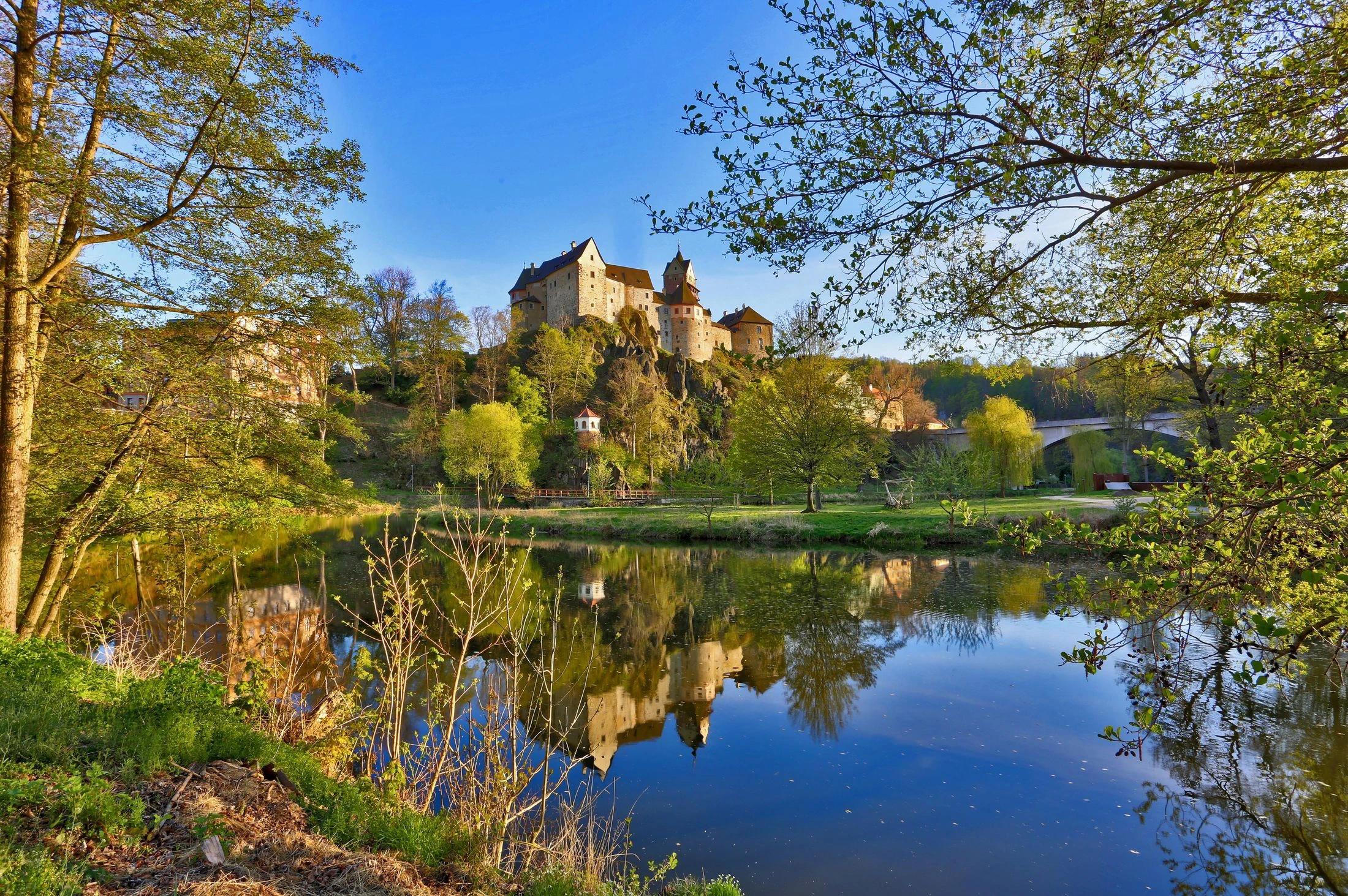 Pohled zpoza jezera na hrad Loket zasazený v malebné krajině.