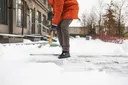 Žena v oranžové zimní bundě odklízí napadaný sníh před vchodem u domu