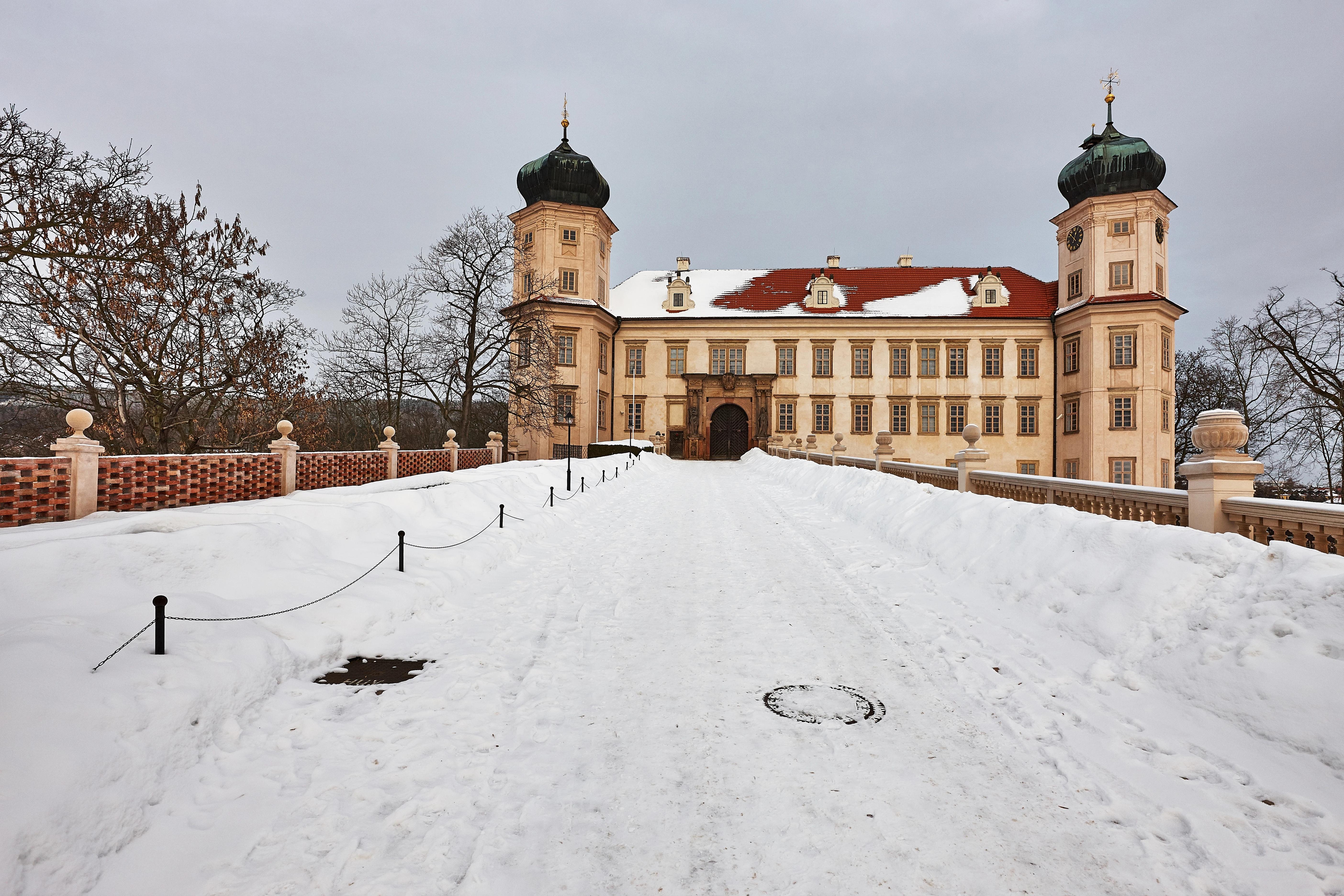 Pohled na zasněženou cestu vedoucí k zámku Mníšek pod Brdy, který má béžovou fasádu, červenou střechou a dvě věže