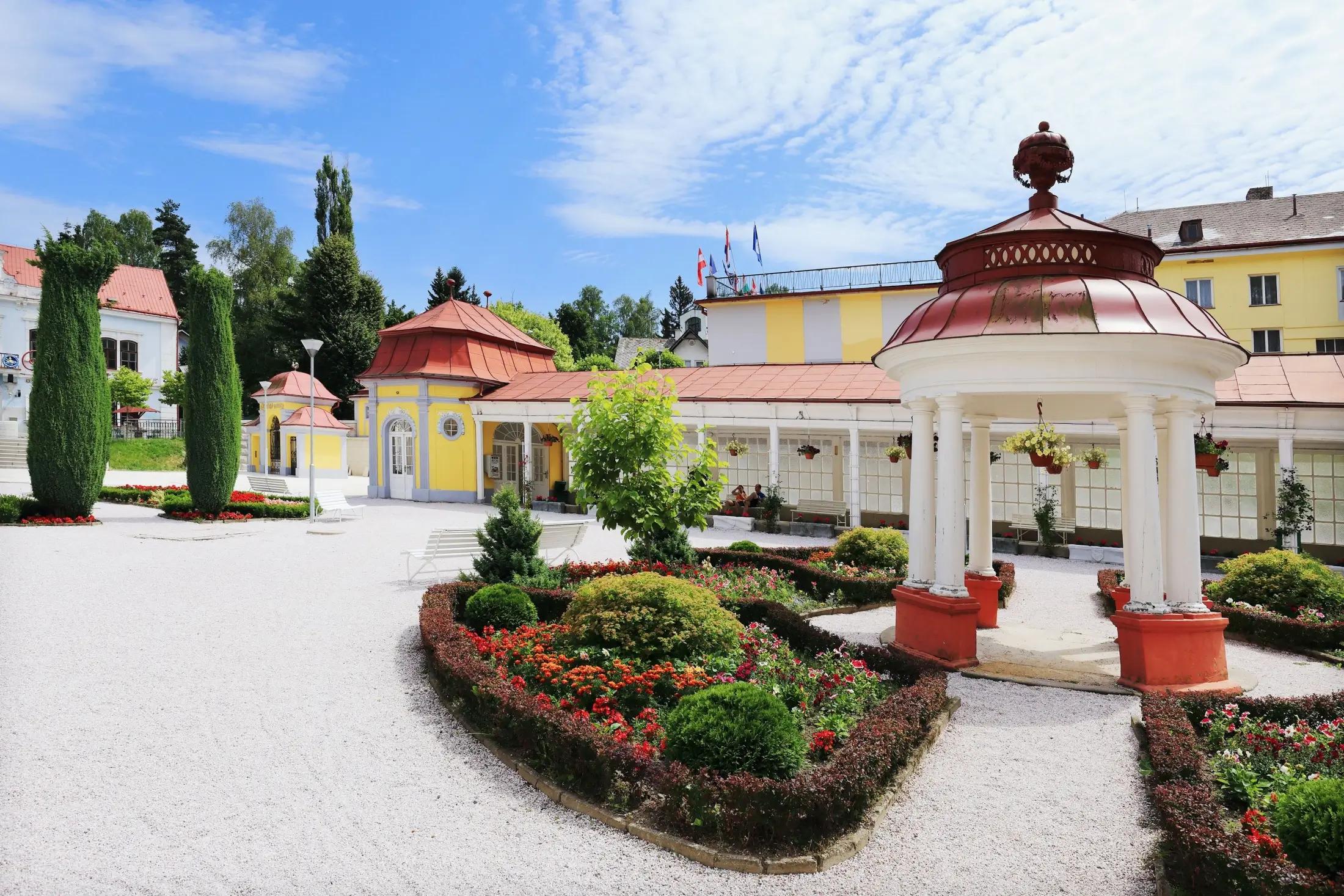 Na obrázku jsou lázně Libverda s krásnými pavilony a květinami. Je krásný den, na obloze je jen pár mraků, jinak je modrá. 