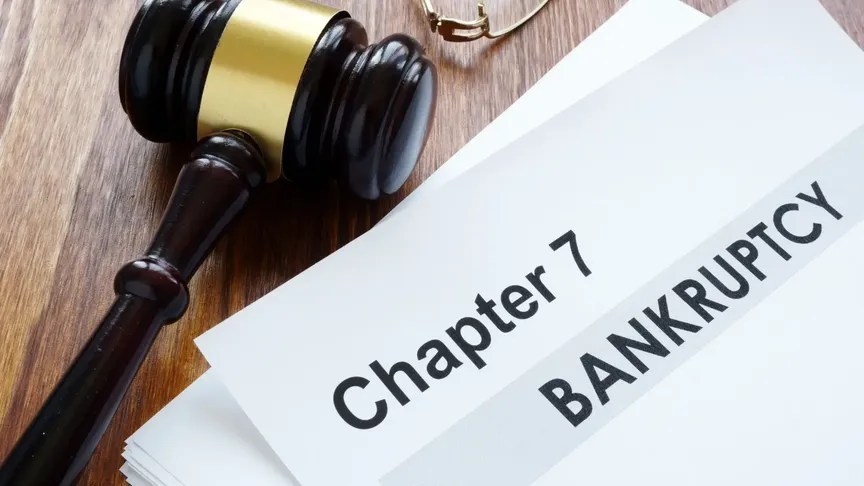 Na hnědém stole leží kladívko soudce a papíry. Na vrchním papíře je napsané „Chapter 7 BANKRUPTCY".