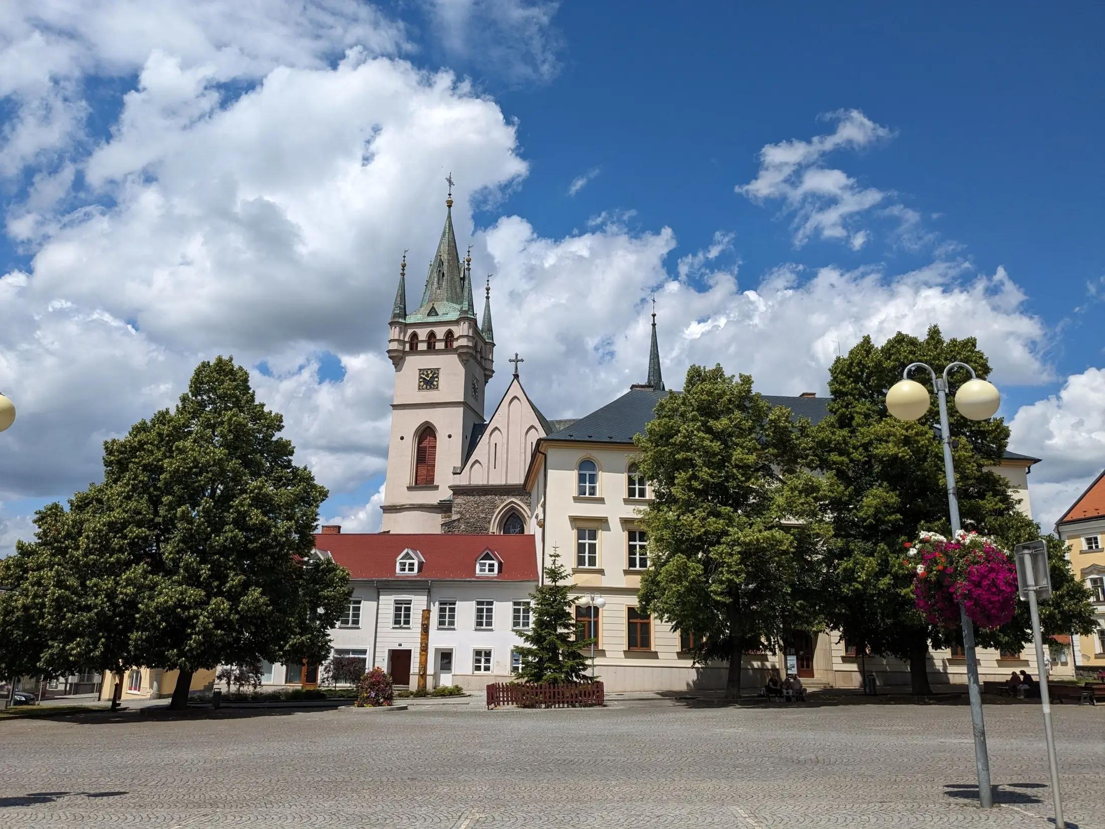 Kostel sv. Mikuláše a statní budovy ve městě Humpolec. Vidět jdou také nějaké zelené stromy a modrá obloha s pár mraky.