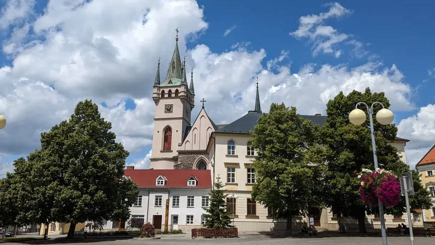 Kostel sv. Mikuláše a statní budovy ve městě Humpolec. Vidět jdou také nějaké zelené stromy a modrá obloha s pár mraky.