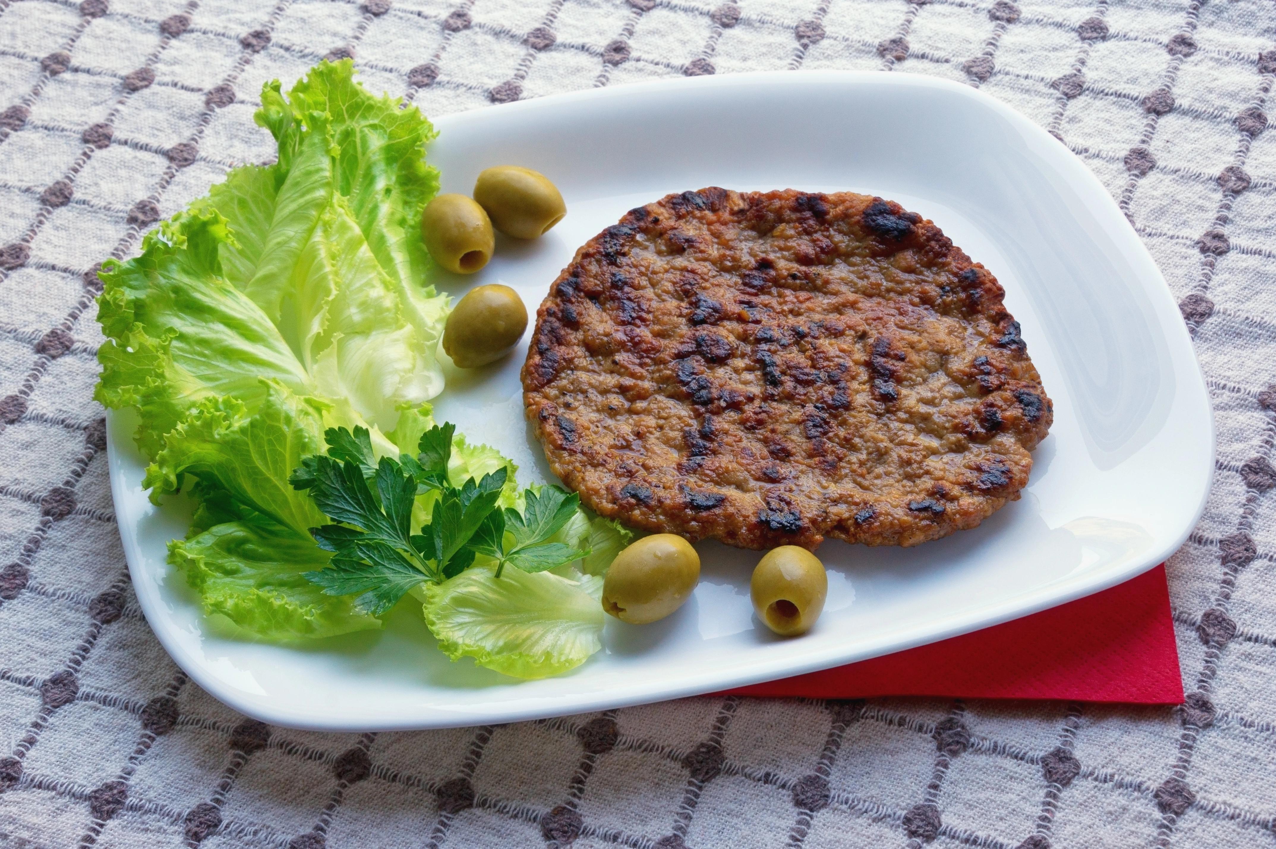 Na bílém talíři se nachází Pljeskavica – grilované jídlo z mletého masa – se zelenými olivami a listy salátu 