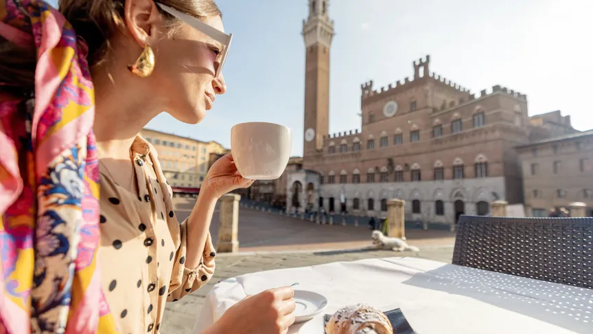Žena snídající italskou snídani s kávou a croissantem, sedící v venkovní kavárně s krásným výhledem na hlavní náměstí města Siena, Itálie.