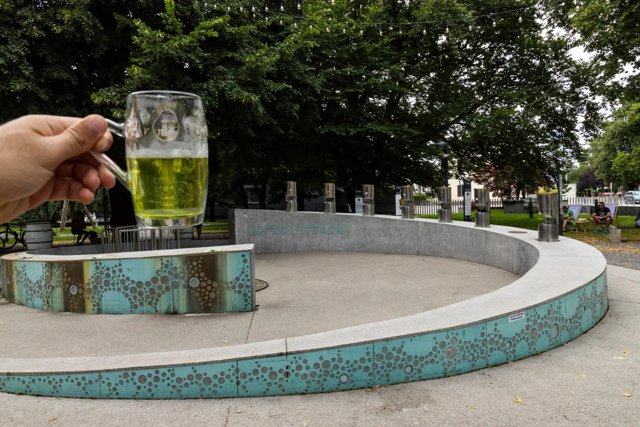 Muž drží v ruce sklenici se zeleným pivem. V pozadí jde vidět pivní fontána.