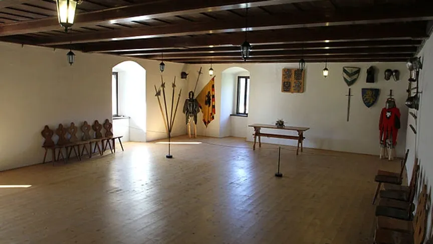 Sál s dřevěnými trámy na stropě, visící lucernou a rytířskou výzdobou