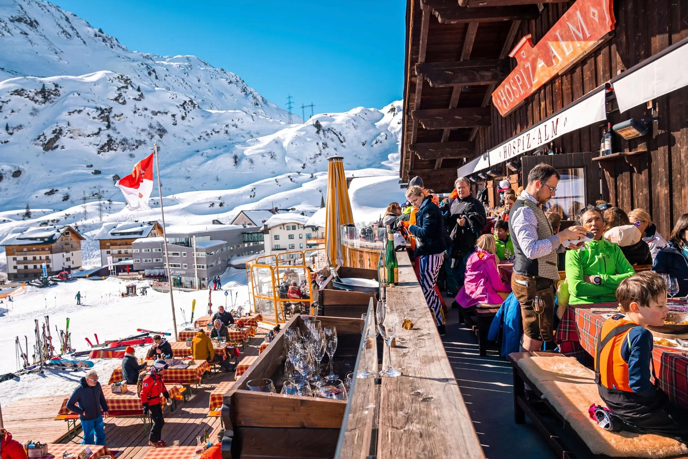 Středisko St. Anton am Arlberg, na pravé straně je horská chata, kde je nejspíše restaurace. Lidé tu sedí na lavičkách u stolů. Na levé straně jsou vidět v dálce další domy a v pozadí hory. Okolí je zasněžené.