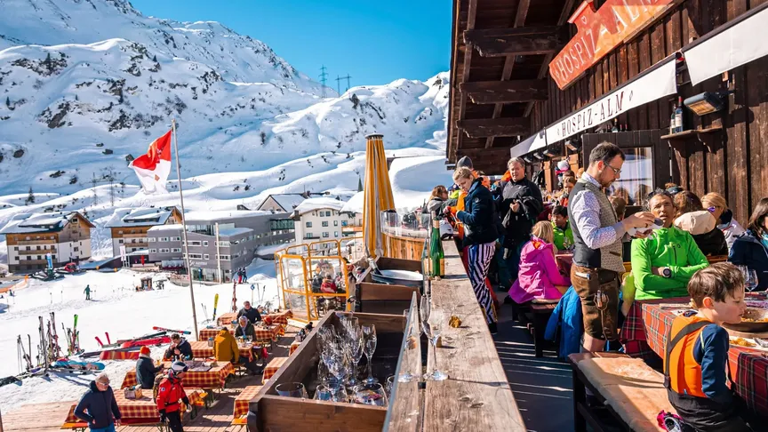 Středisko St. Anton am Arlberg, na pravé straně je horská chata, kde je nejspíše restaurace. Lidé tu sedí na lavičkách u stolů. Na levé straně jsou vidět v dálce další domy a v pozadí hory. Okolí je zasněžené.