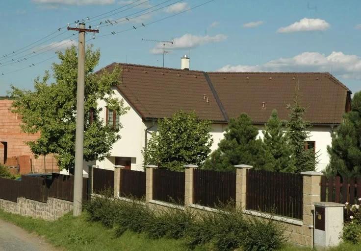 Hercova bílá chalupa s hnědou střechou, která je schována za dřevěný plot a část jehličnatých stromů