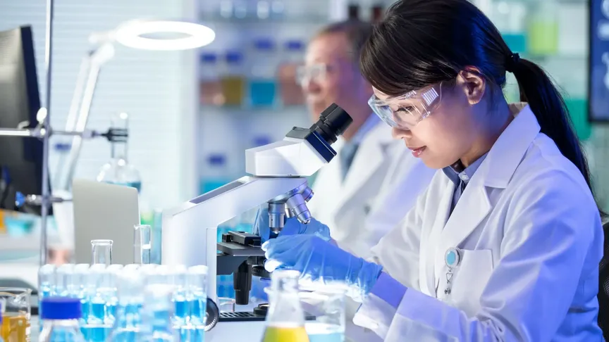 Obrázek vědců v laboratoři. Více v popředí sedí mladá žena u stolu, kde jsou zkumavky a mikroskop. Má na sobě bílý plášť a ochranné brýle.