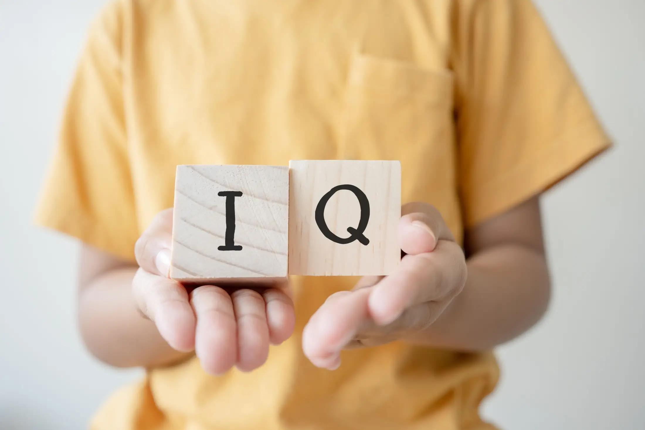 Menší dítě drží dvě kostky, kde jsou písmenka I a Q. Dítěti jde vidět jen torso, ne hlava ani spodní část těla. Má na sobě žluté tričko.