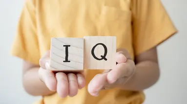 Menší dítě drží dvě kostky, kde jsou písmenka I a Q. Dítěti jde vidět jen torso, ne hlava ani spodní část těla. Má na sobě žluté tričko.