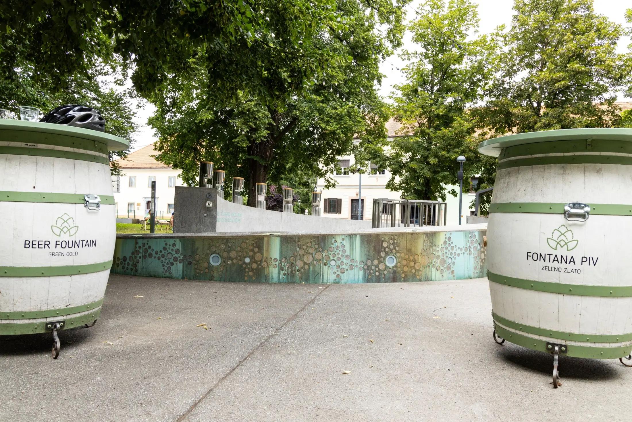 Fotka pivní fontány v zeleném parčíku. Vidět jsou i dva bílo-zelené sudy, které slouží jako stoly.