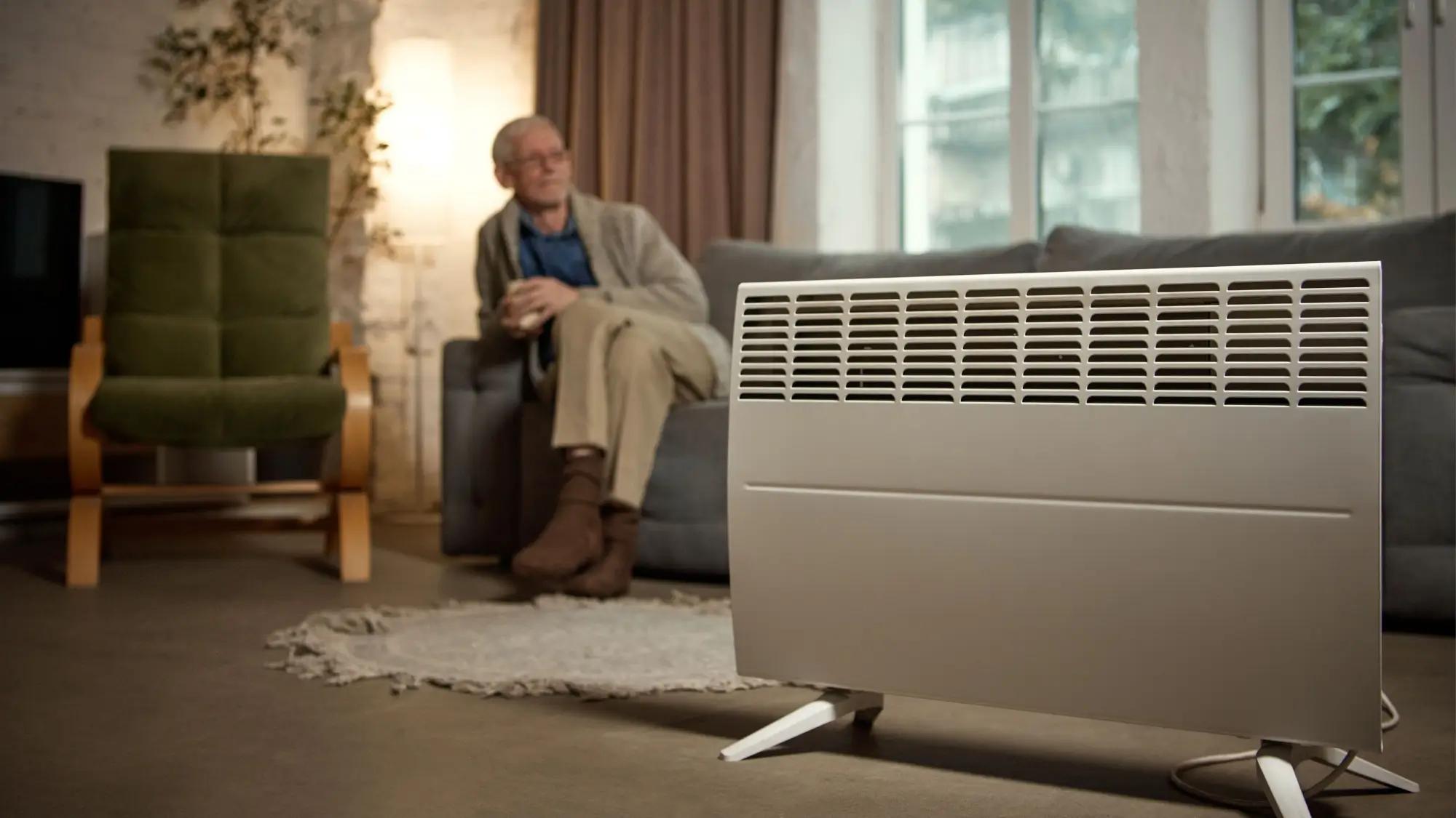 Muž sedí na gauči v teplém oblečení, v popředí je topení