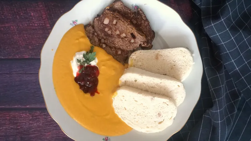 Na tmavém dřevěném stole leží bílý talíř se svíčkovou omáčkou, masem a třemi knedlíky.