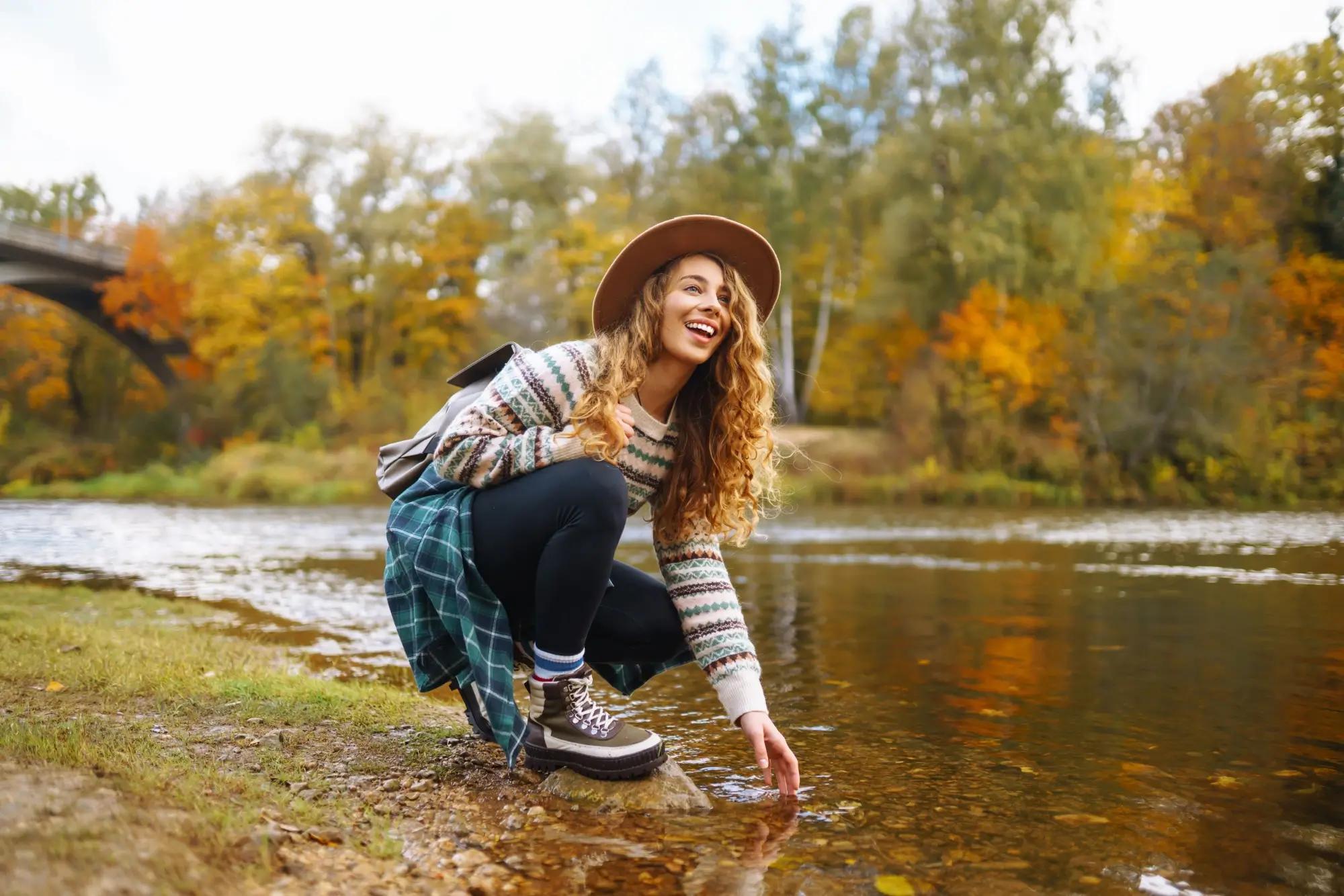 mladá žena zkouší teplotu řeky, za ní krásně zbarvený lesík 