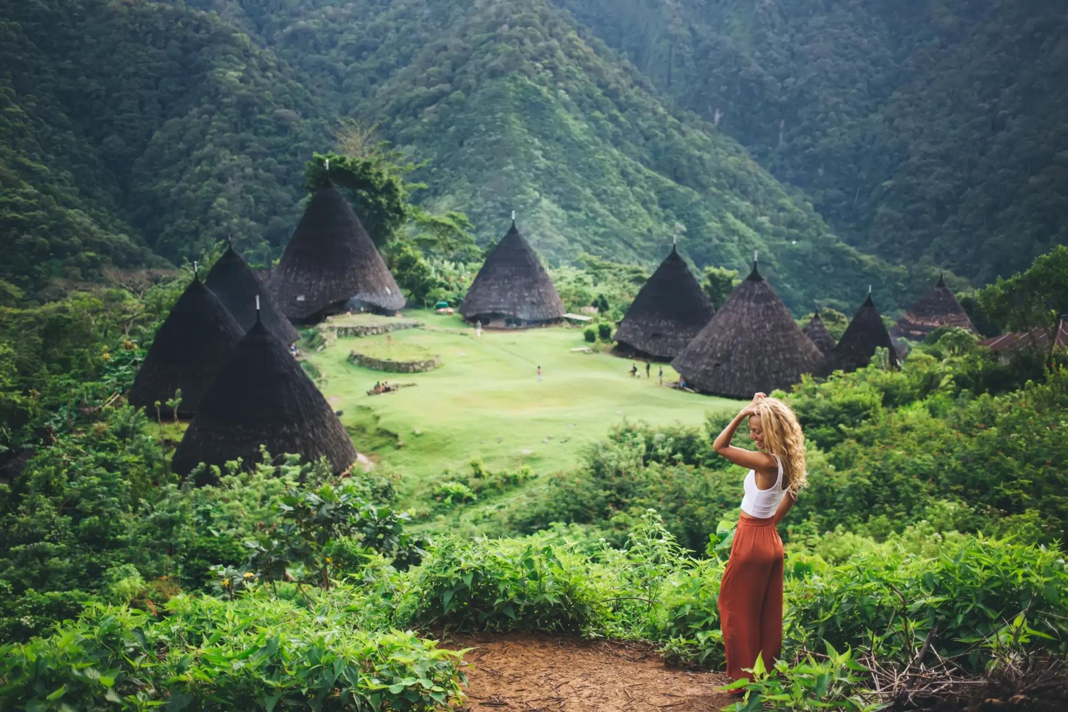 Záběr na chýše vesnice Wae Rebo. Vidět jde také zelené okolí. V popředí stojí mladší žena, pózující pro fotografii.