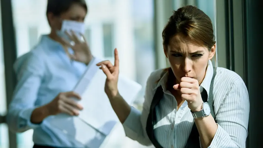 žena v práci kašle, za ní stojí asijská kolegyně s rouškou, která se chrání rukou