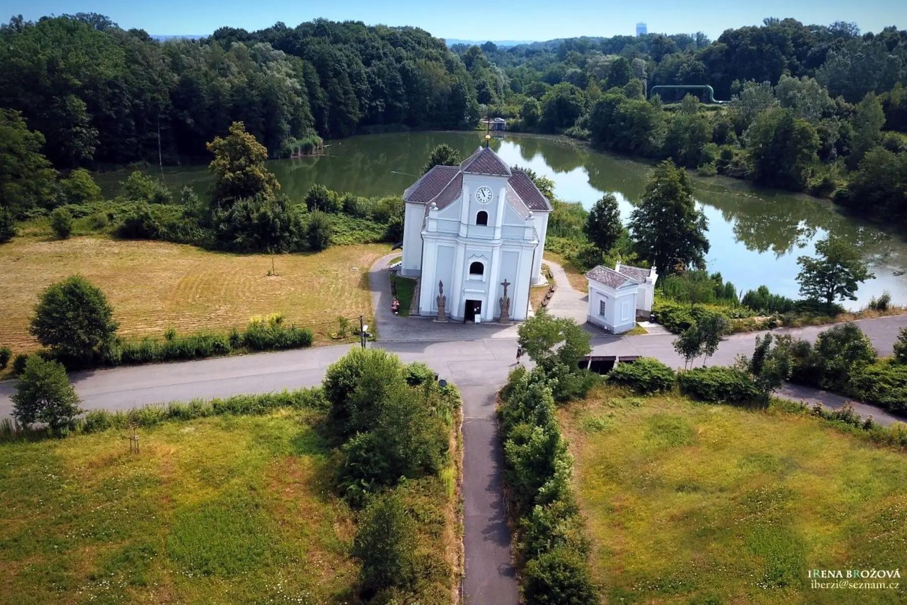 Letecký záběr na kostel sv. Petra z Alkantary. Vedou k němu čtyři cesty, za ním je rybník/nějaká vodní plocha a v pozadí ještě les.