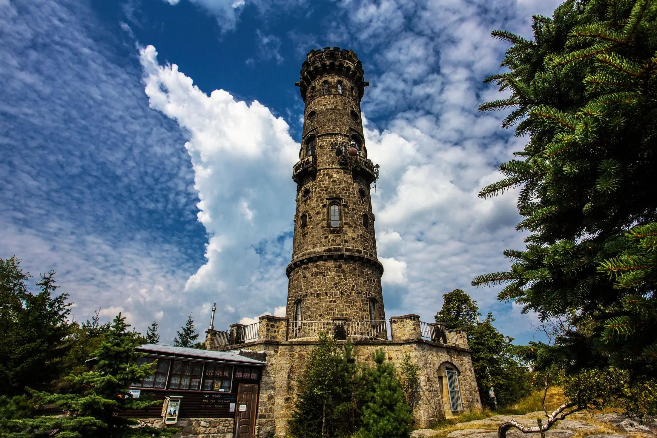 Kamenná rozhledna Děčínský Sněžník za krásného dne. Okolí je plné zelených stromů a obloha modrá s bílými mraky.