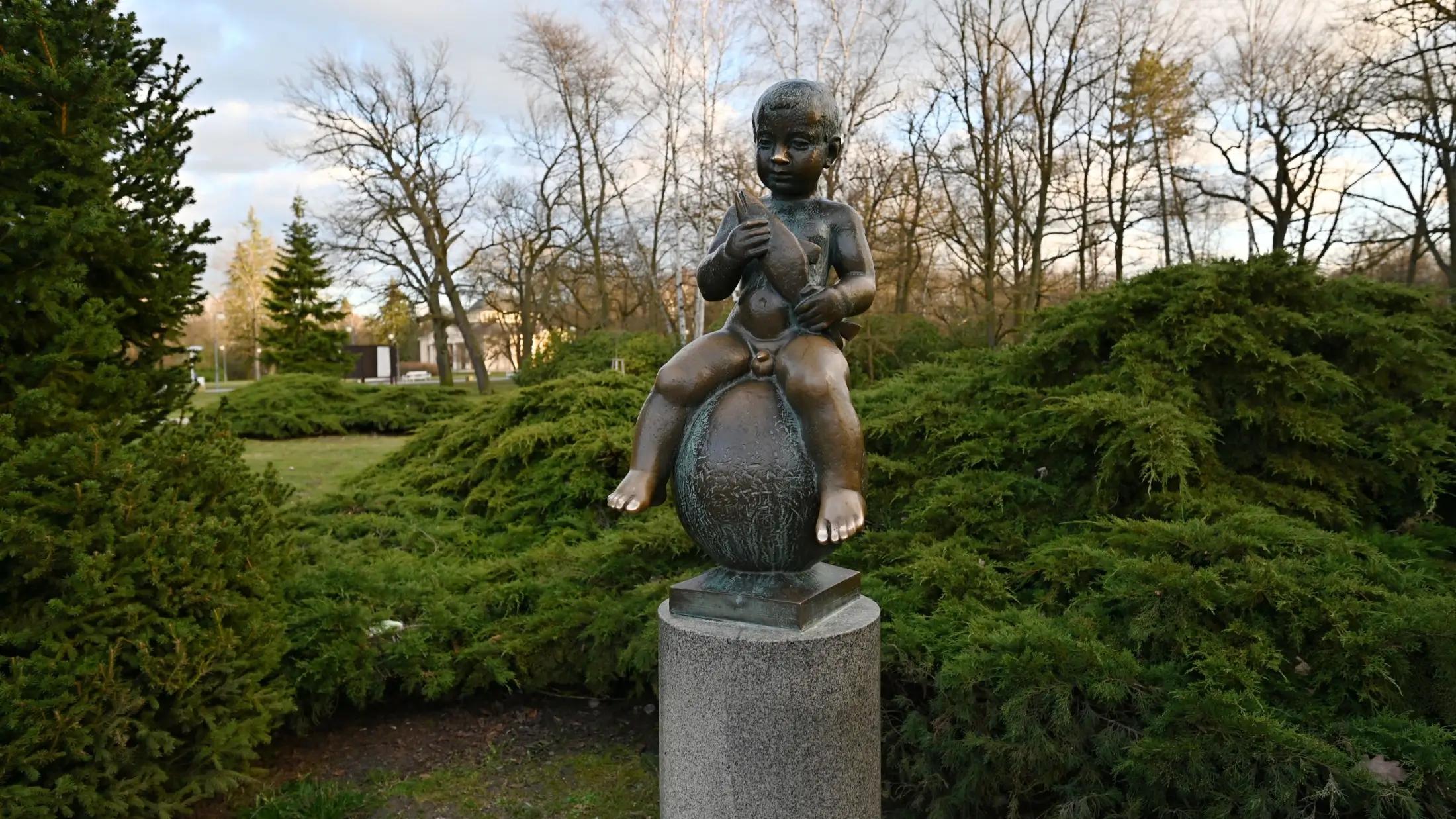 Plastika malého chlapce s rybou. Je podzim, v pozadí jsou stromy buď se žlutými nebo s opadaným listím.