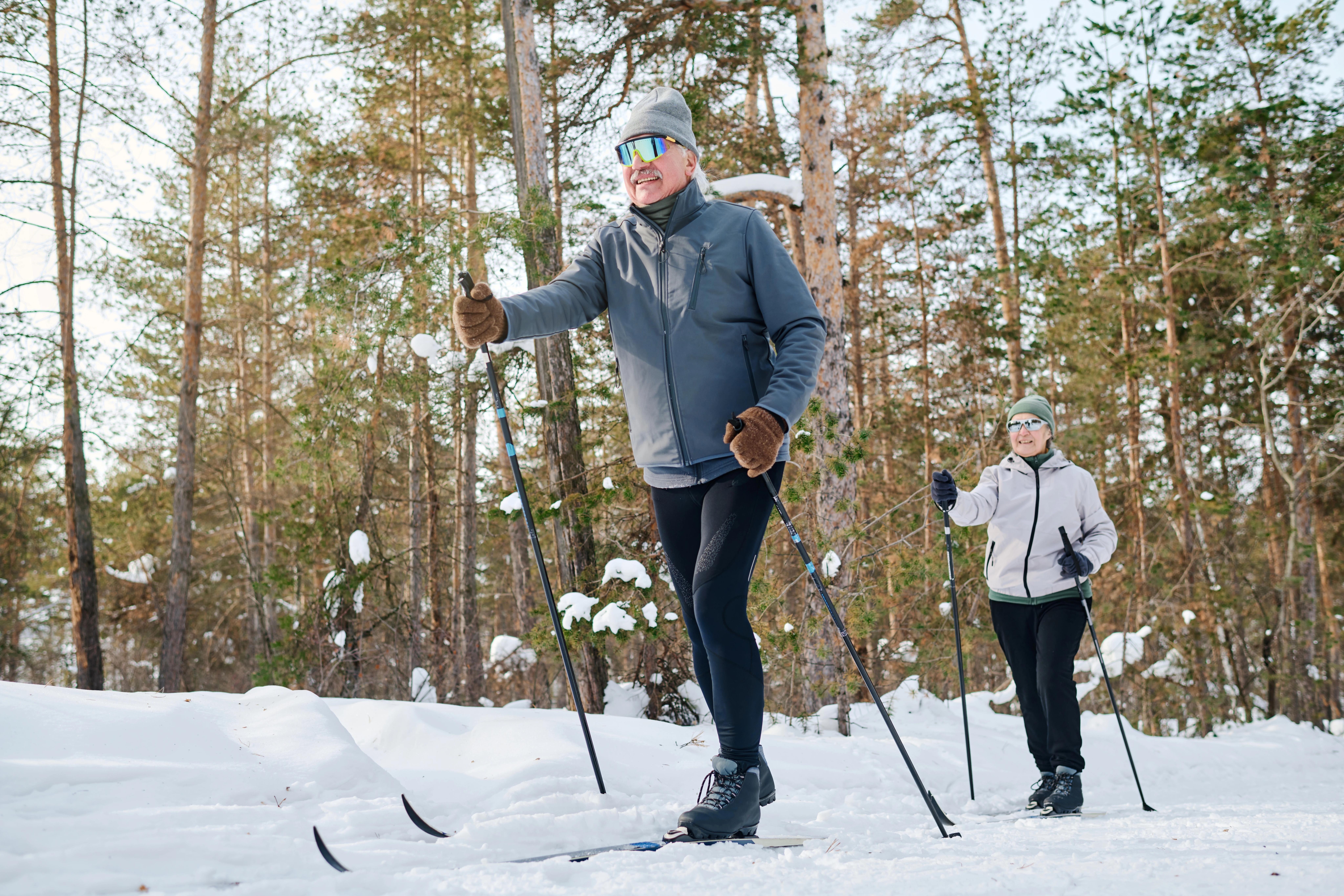 V popředí je starší muž na běžkách, za ním je starší žena na běžkách. V pozadí jsou stromy