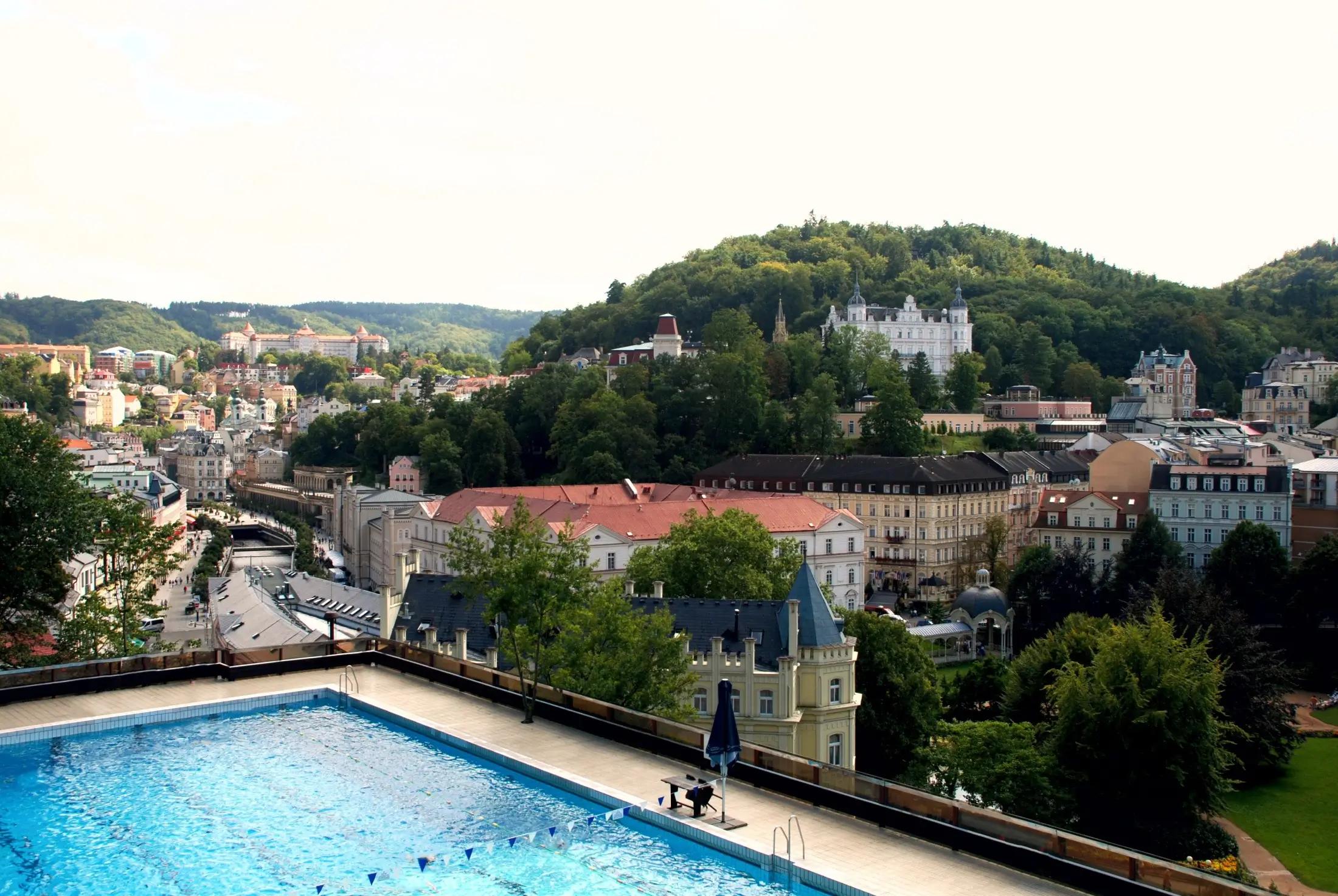 Panoramatický pohled na část střešního bazénu hotelu Thermal a okolí, které ze střechy je možné vidět. Vidět jsou unikátní budovy a zeleň stromů.