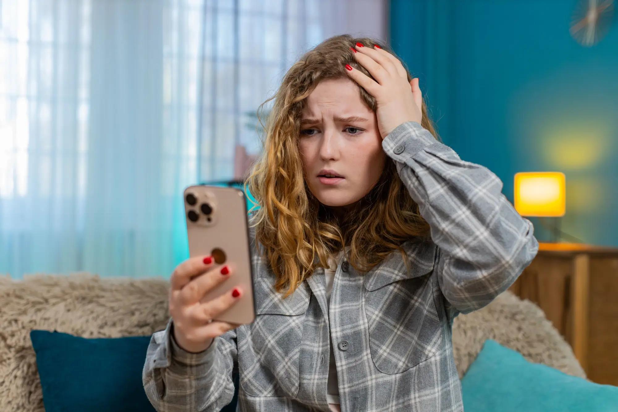 Rozrušená mladá žena se dívá do telefonu a drží se za hlavu