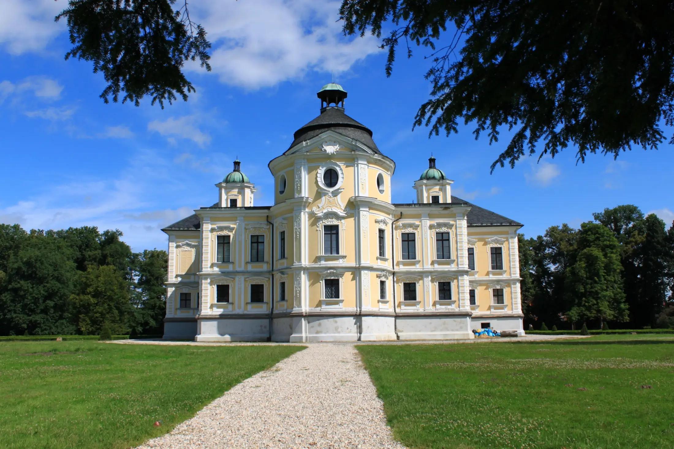 Zámek Kravaře s cestou, nejspíše z kamínků, vedoucí k němu. Je krásný den, modrá obloha s pár mraky. Okolí je plné zeleně.