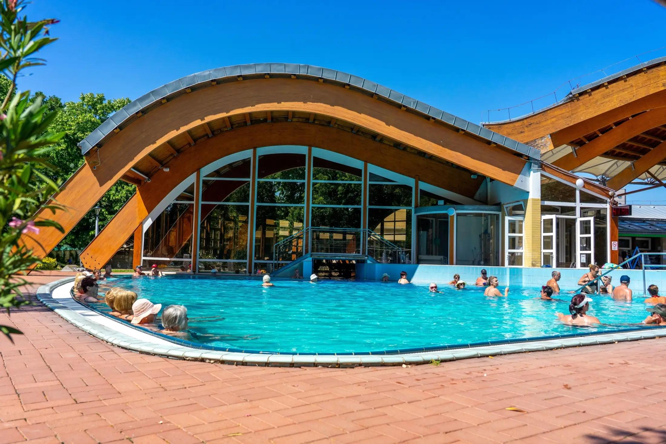 Fotka lázní Bükfürdő Thermal & Spa. Vidět jde budova a před ní jeden z venkovních bazénů, ve kterém je pár lidí. Je krásný den, svítí Slune a obloha je bez mráčku.