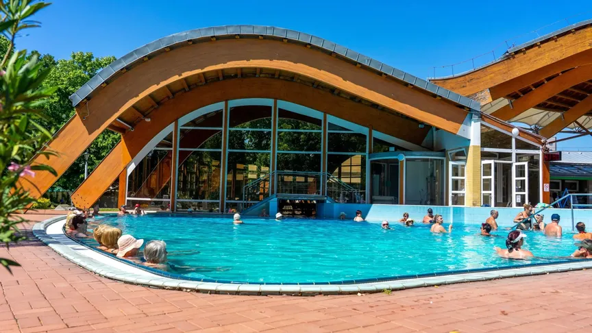 Fotka lázní Bükfürdő Thermal & Spa. Vidět jde budova a před ní jeden z venkovních bazénů, ve kterém je pár lidí. Je krásný den, svítí Slune a obloha je bez mráčku.