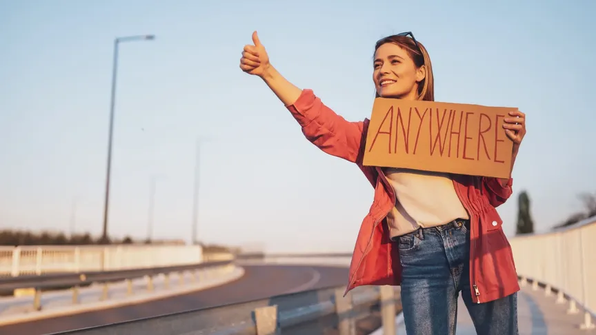 Mladá žena stojí u silnici. Má na sobě modré džíny, bílou halenku a červenou bundu. Stopuje, protože drží palec pravé ruky nahoře a v druhé ruce má ceduli s anglickým slovem "anywhere", česky přeloženo "kamkoli".