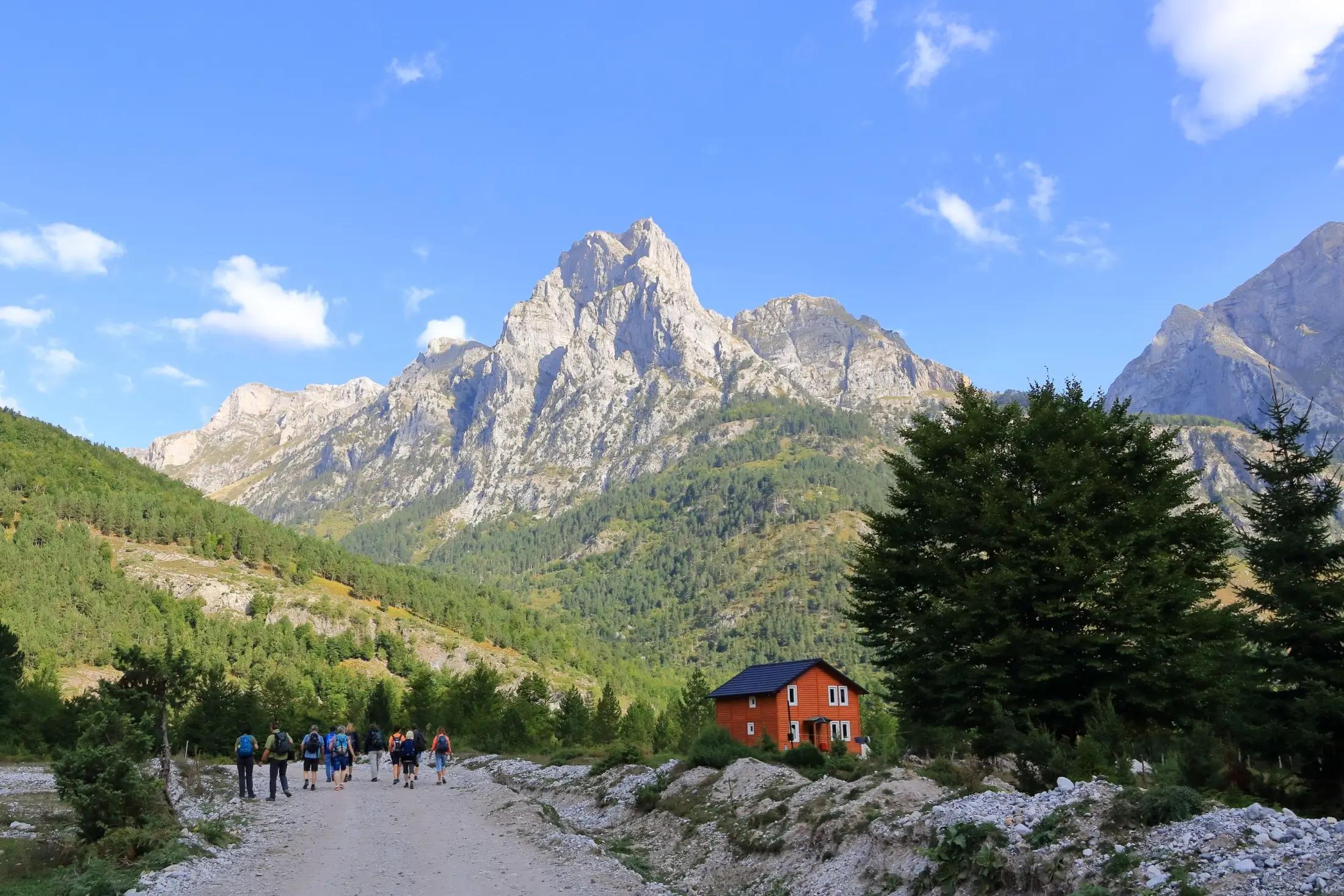 Záběr na hory tyčící se do modrého nebe. Okolí je poseto množstvím stromů. Uprostřed obrázku jde vidět červený domek a nalevo od něj v dáli je parta turistů, kteří jdou na túru.