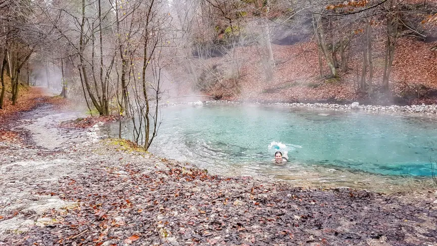 Přírodní pramen Maibachl s průzračnou modrou vodou. Je podzim nebo zima, na stromech okolo není žádné listí. V prameni se koupe mladá žena.