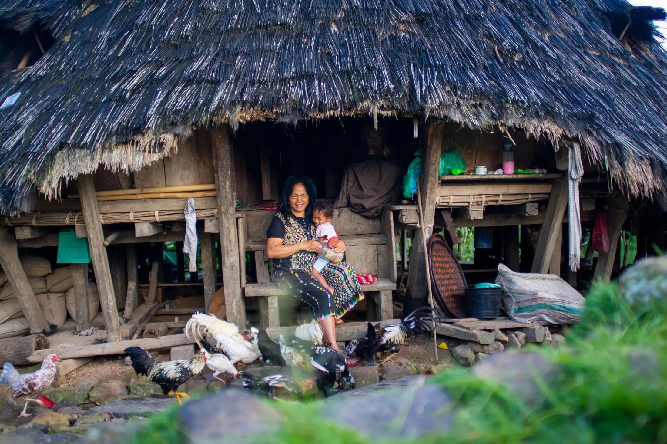 Jedna žena z vesnice Wae Rebo sedí na schodech chýše. Na klíně drží malé dítě.