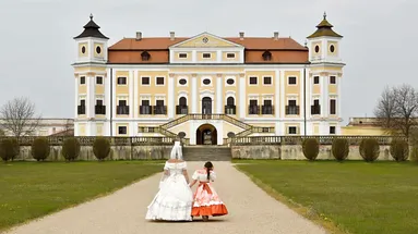 žena s holčičkou v historickém kostýmu se prochází po zámecké zahradě