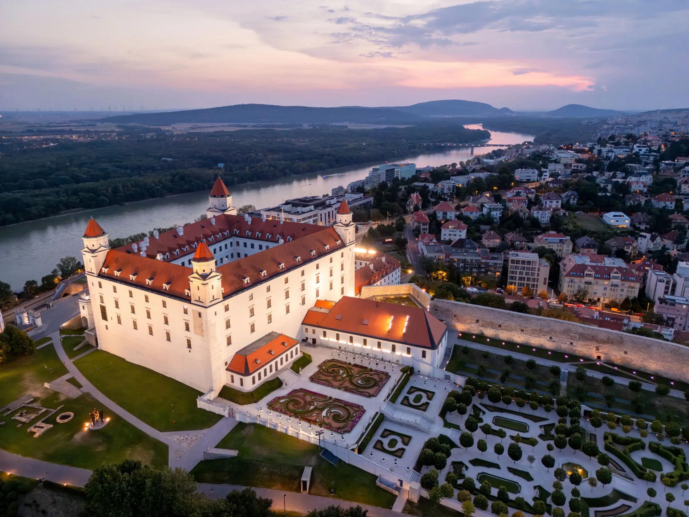 Letecký pohled na Bratislavský hrad a jeho zahrady za soumraku. Vidět jde také řeka Dunaj a na pravé straně ostatní domy města.