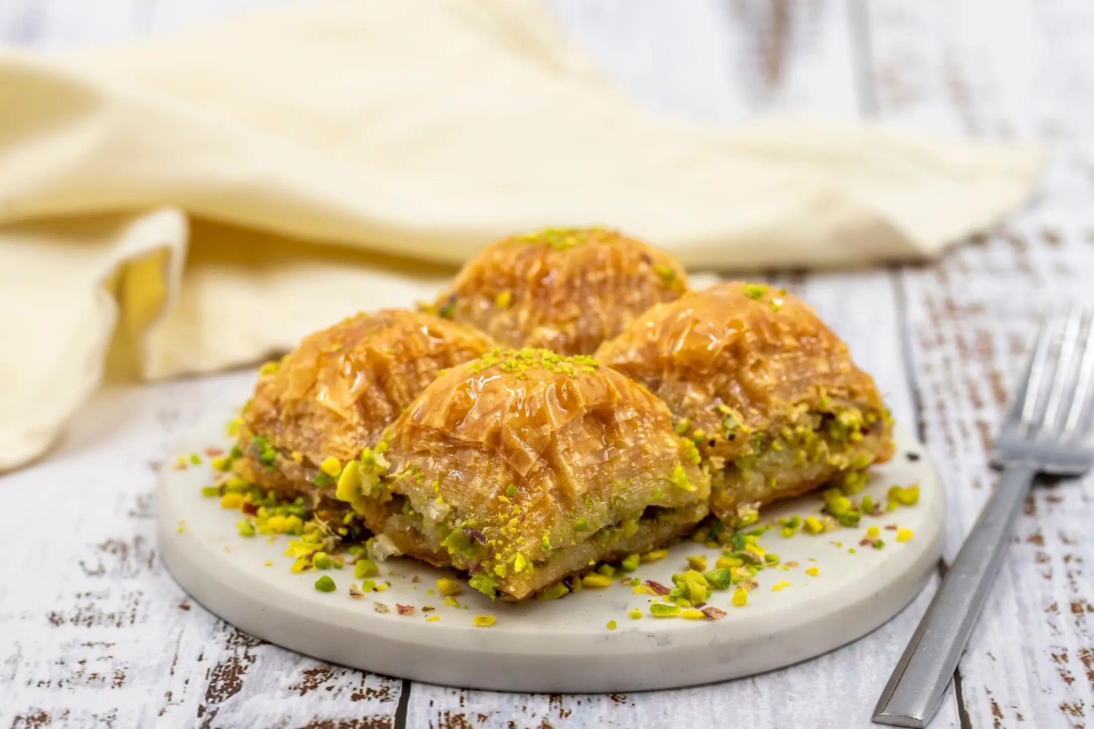 Baklava ležící na menším šedém talířku. Vidět jde stříbrná vidlička na pravé straně fotky a béžový ubrousek v pozadí.