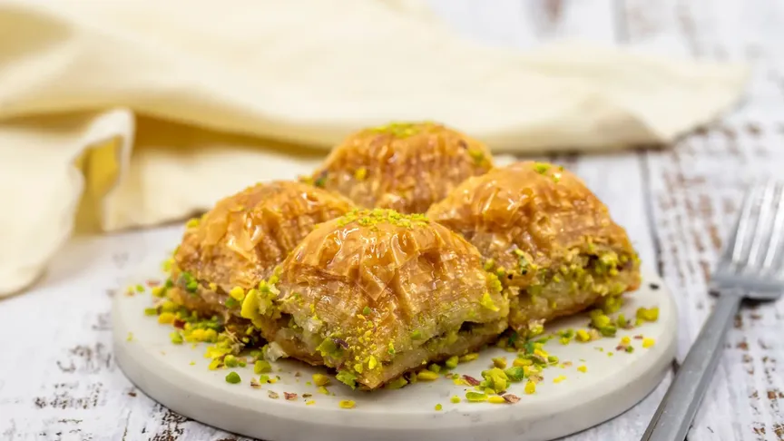 Baklava ležící na menším šedém talířku. Vidět jde stříbrná vidlička na pravé straně fotky a béžový ubrousek v pozadí.