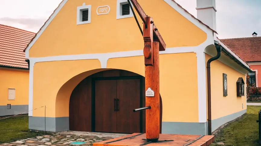 Menší žlutá budova ve stylu selského baroka. Před ní stojí něco, co vypadá jako starodávná pumpa na vodu.