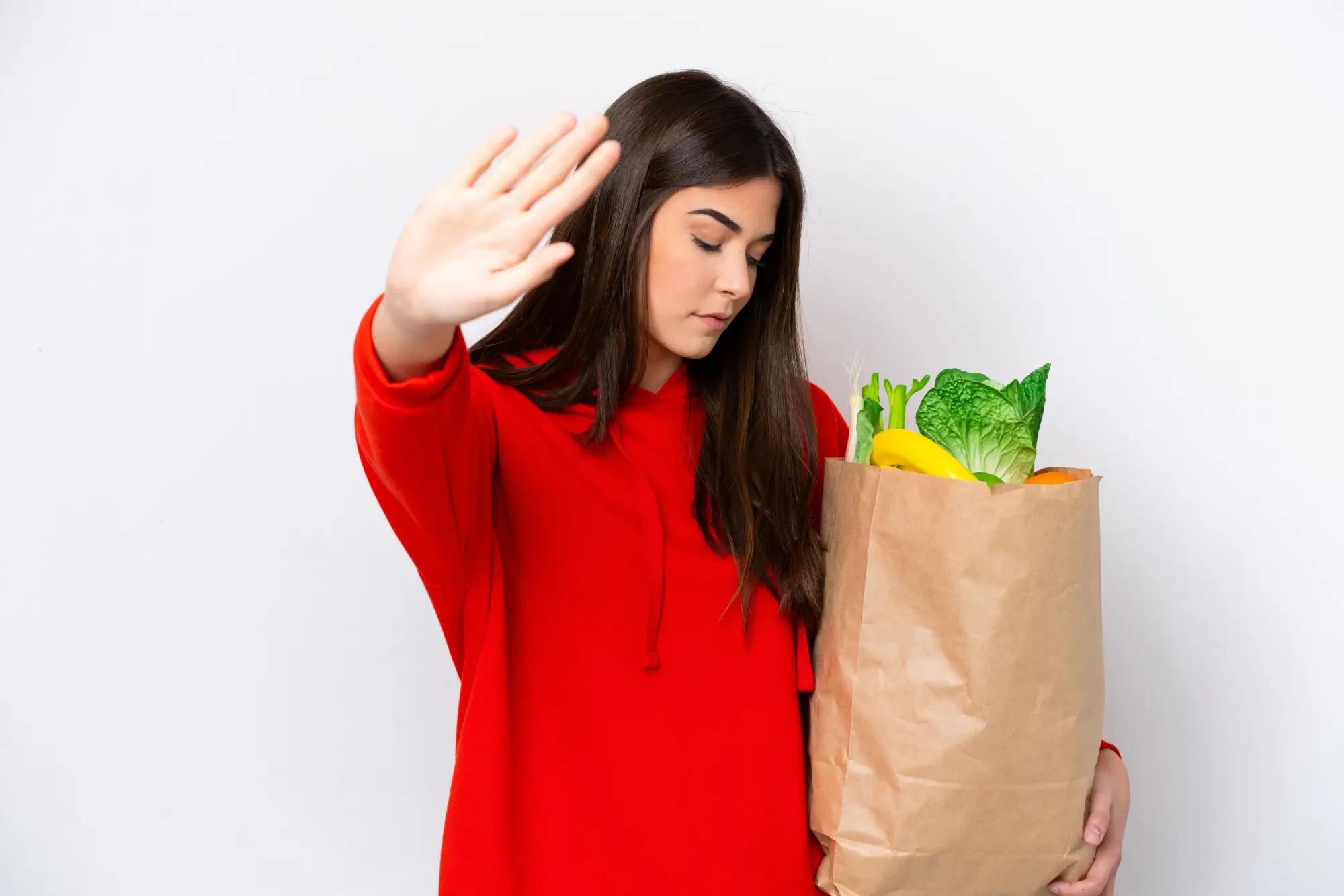 Obrázek ukazuje ženu v červené mikině, která drží tašku s potravinami. Má ruku nahoře v odmítavém gestu.
