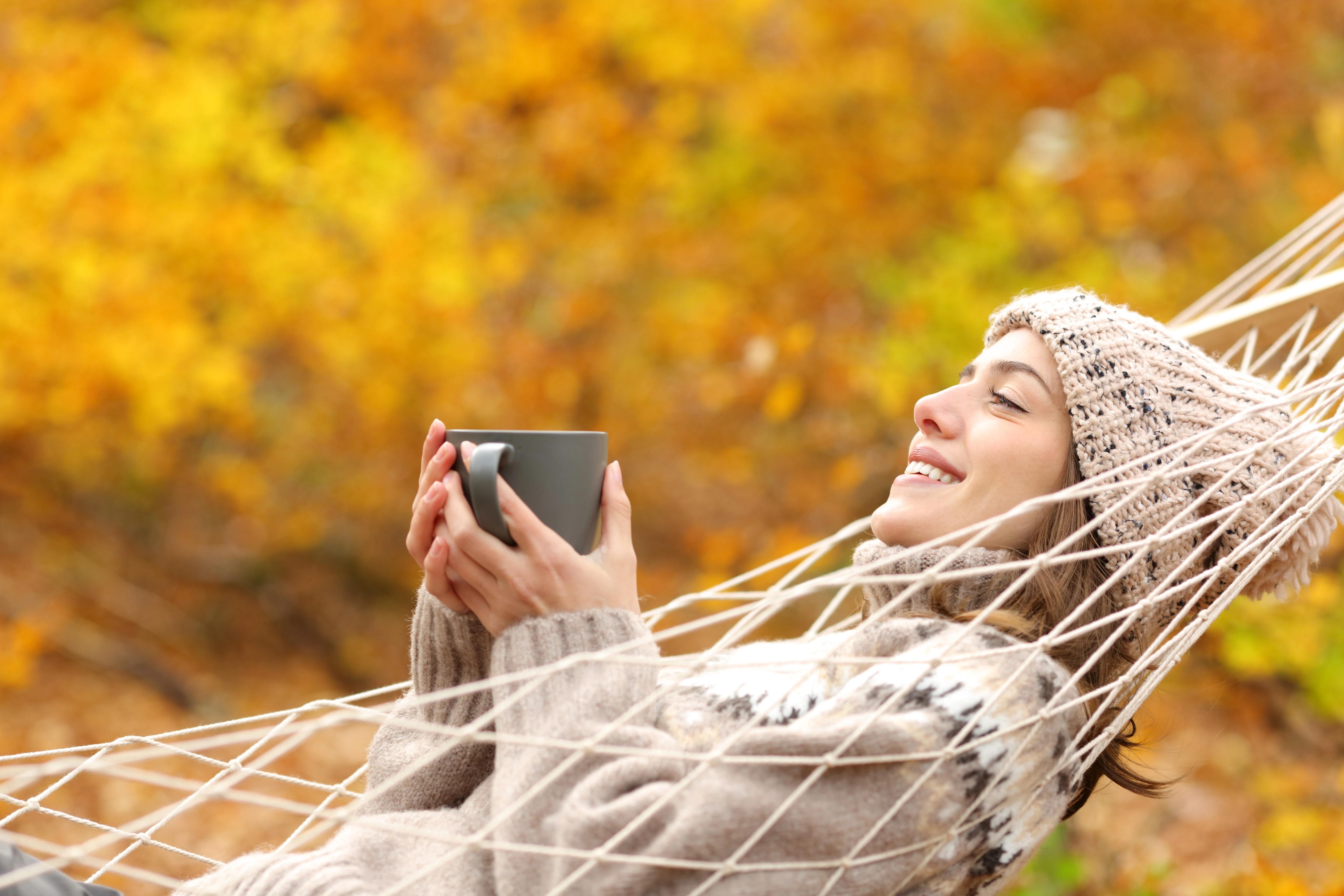 Smějící se žena v béžové čepici a svetru si v houpací síti užívá svůj čaj ve slunečném podzimním dni