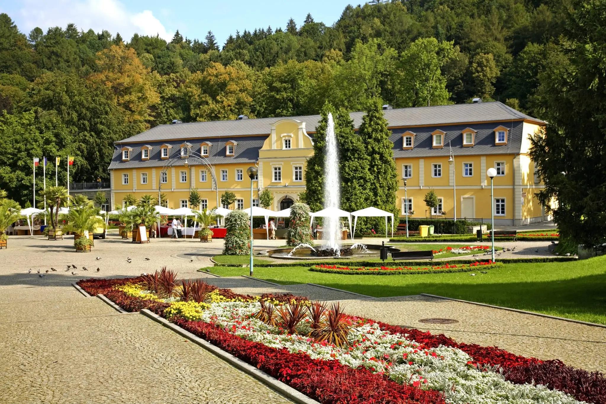 Zelené stromy se týčí nad lázněmi a lázeňskou kolonádou s tryskající fontánou v Kudowa-Zdrój