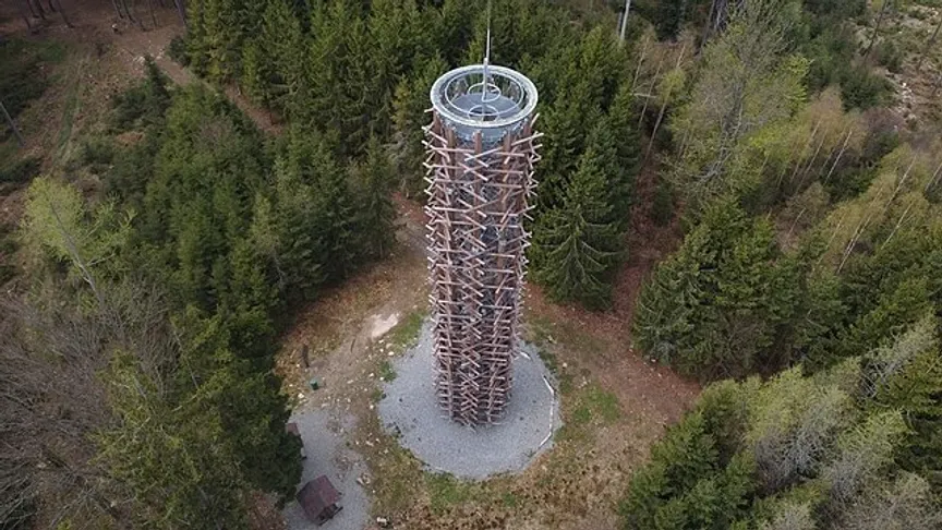 Uprostřed zelenajících se lesů na kulatém plácku, stojí rozhledna obložená dřevěnými trámy s kovovou konstrukci