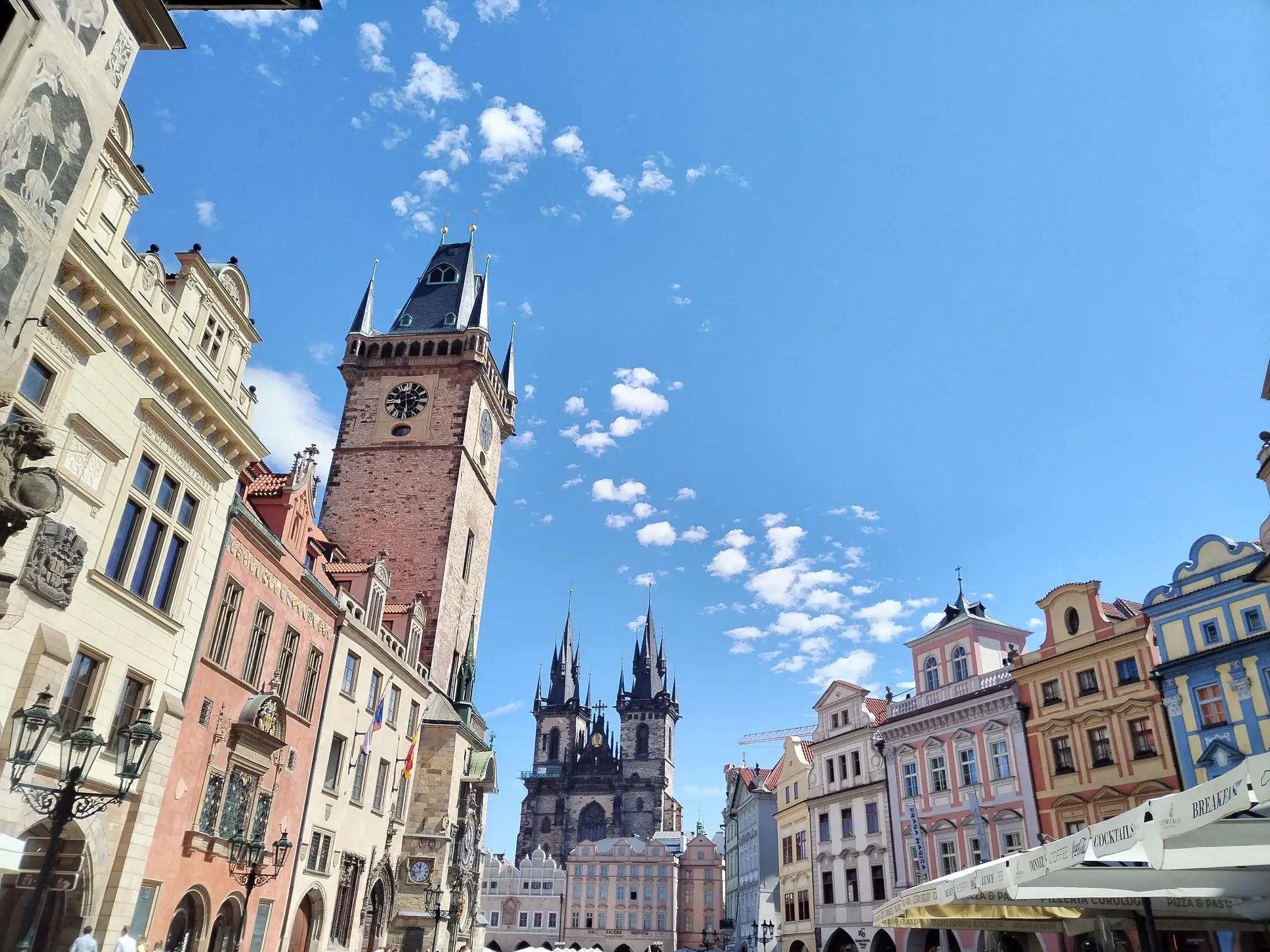 Staroměstké náměstí v Praze za krásného jasného dne. Vidět jde nalevo radnice s orlojem. V prostředku chrám Matky Boží před Týnem.