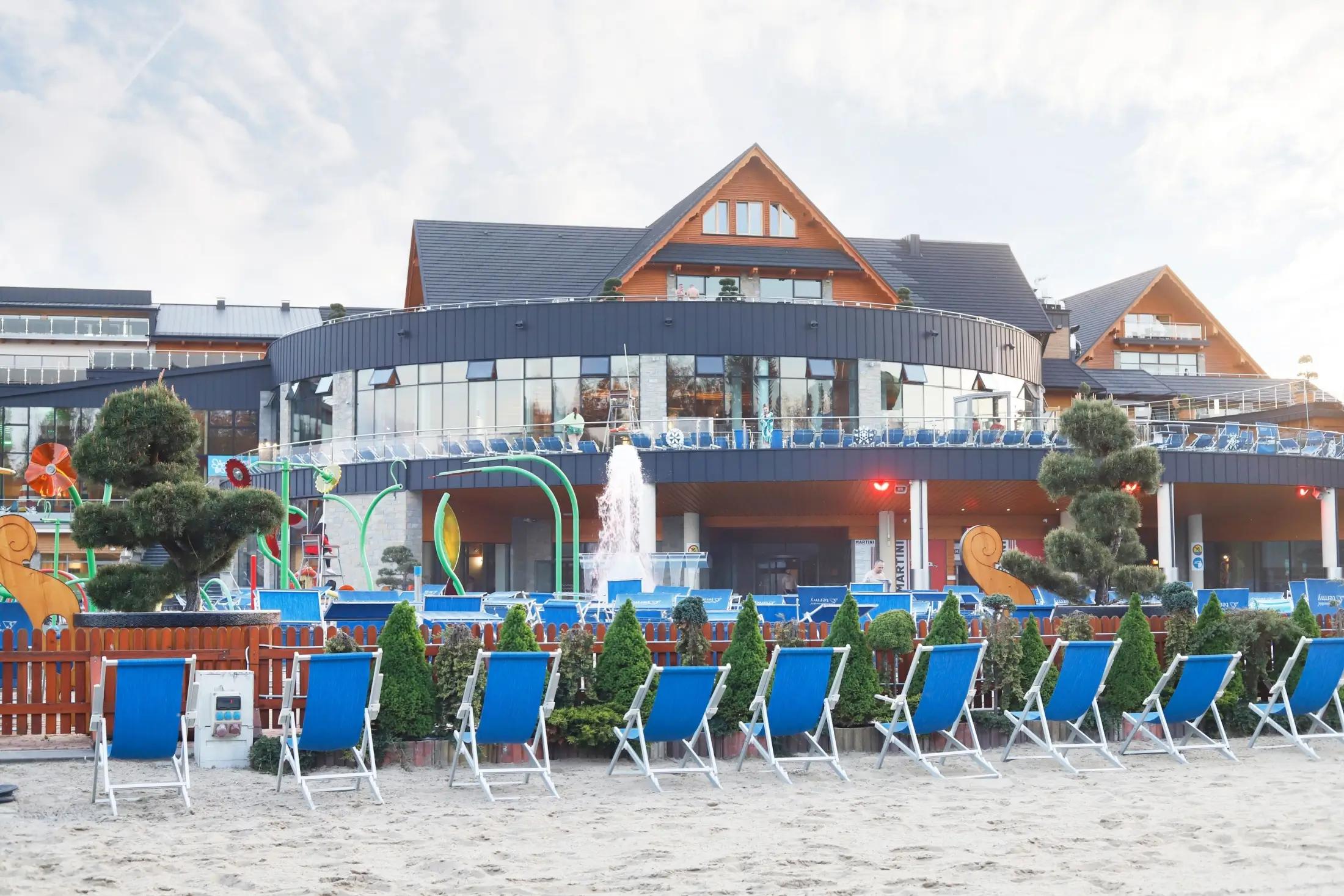 Obrázek ukazuje zevnějšek lázní, jde vidět hlavní budova a před ní písečná pláž a lehátka.