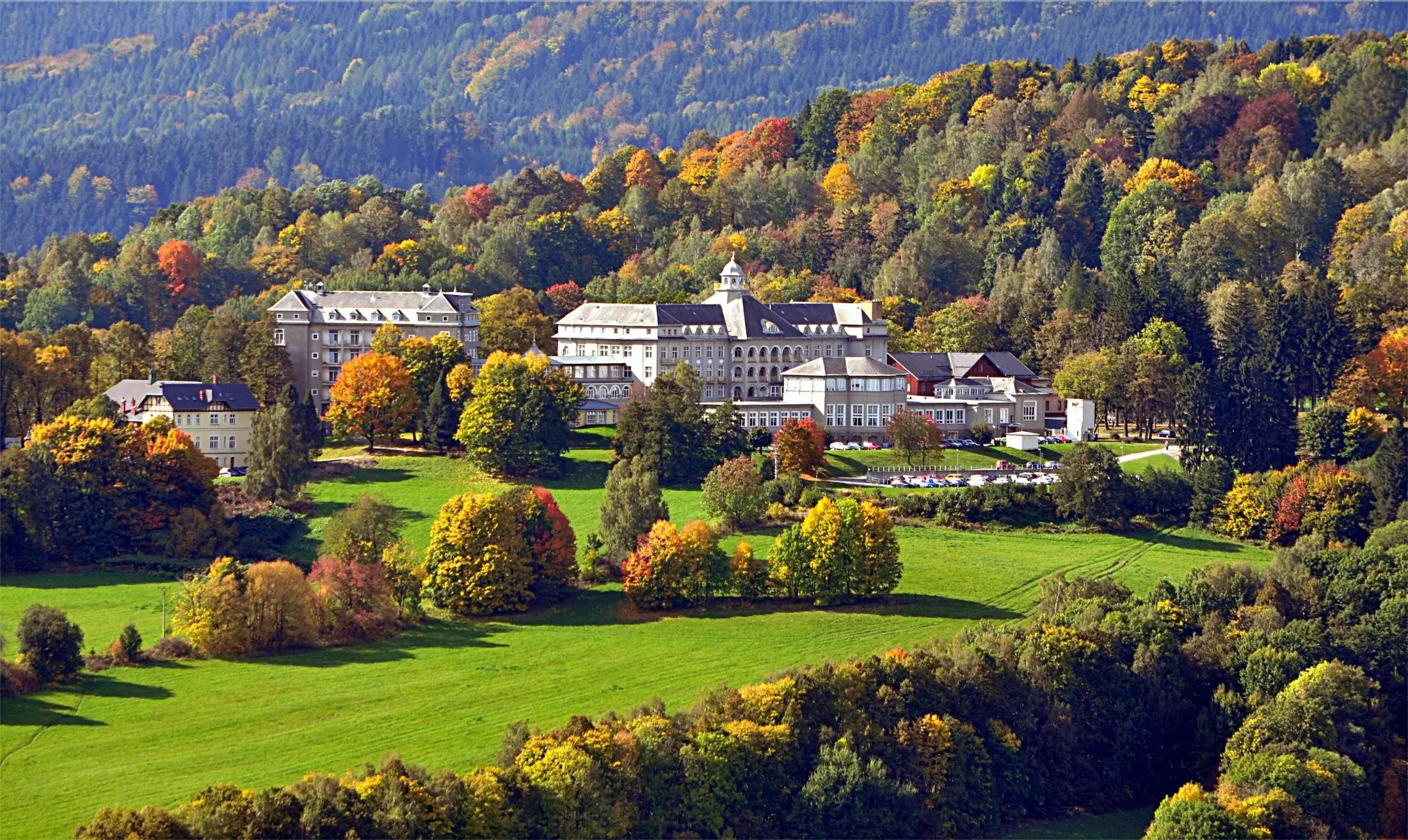 Panoramatický záběr na Priessnitzovy lázně a jejich okolí. Je podzim, jelikož okolní stromy se začínají lehce zbarvovat do žluté a červené barvy. 