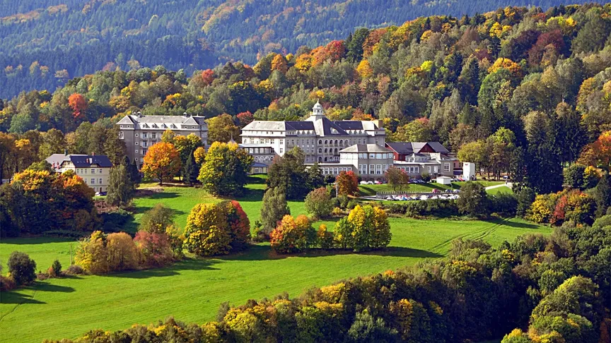 Panoramatický záběr na Priessnitzovy lázně a jejich okolí. Je podzim, jelikož okolní stromy se začínají lehce zbarvovat do žluté a červené barvy.