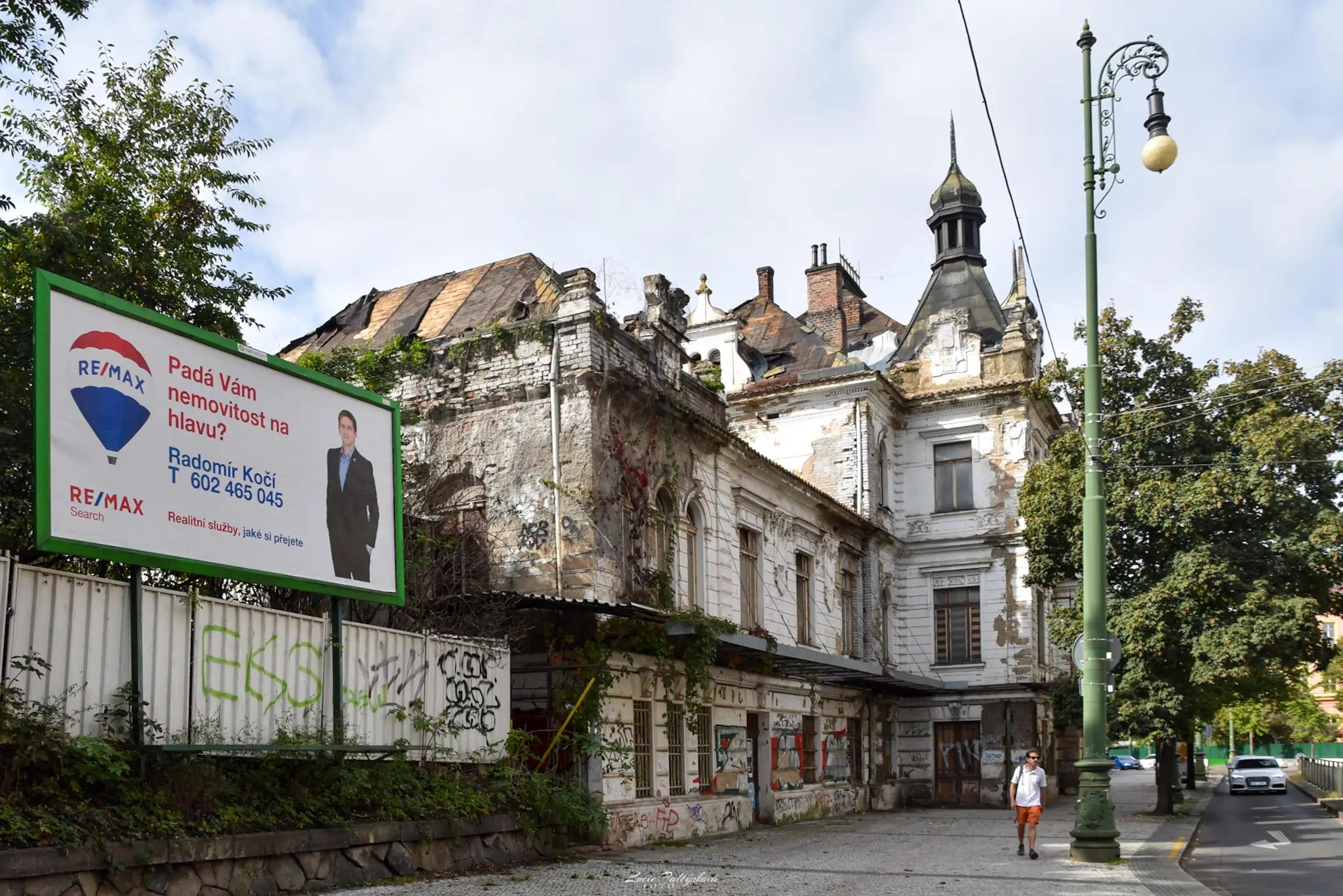 Pohled na rozpadající se nádražní budovu s billboardem realitní kanceláře s nápisem: Padá vám nemovitost na hlavu?